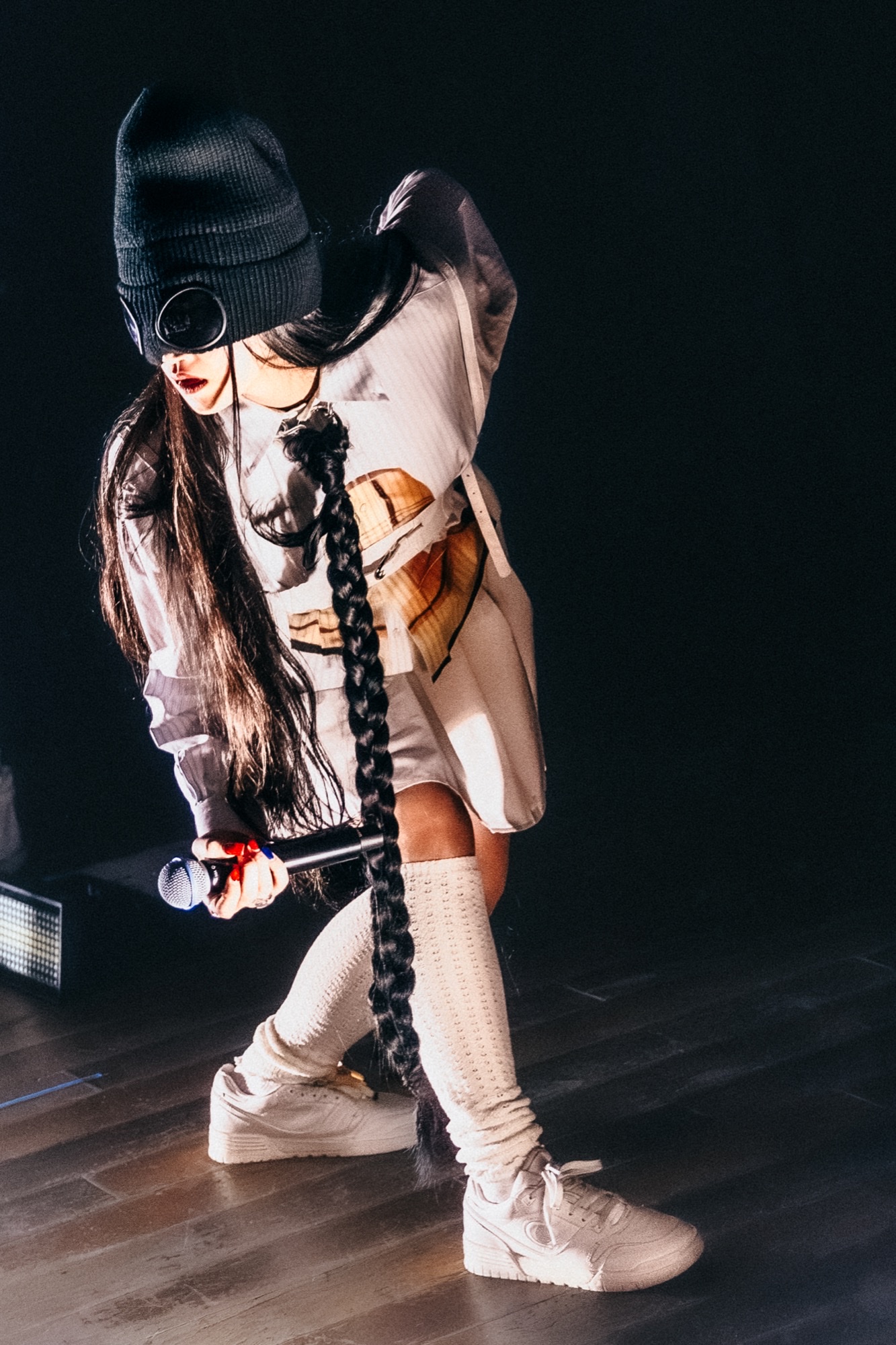 Audrey Nuna crouched onstage in a beanie and graphic shirt, mic in hand under a hard spotlight.