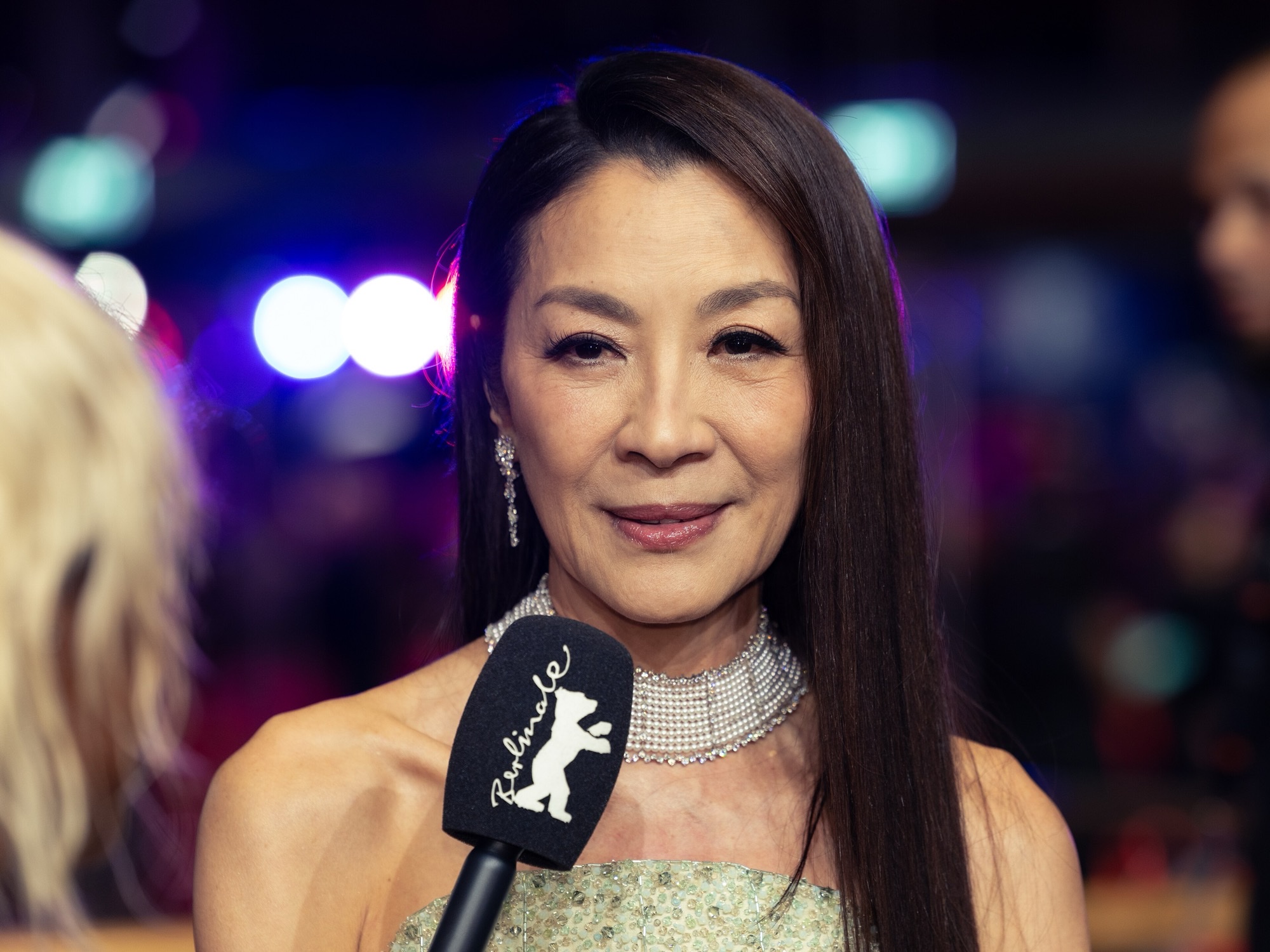 Michelle Yeoh at the 2026 Berlin Film Festival, in a pale green strapless gown and pearl choker, speaking into a Berlinale microphone.